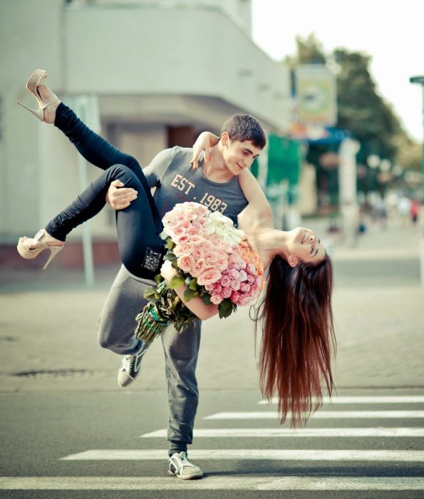 Chico cargando a una chica que trae en sus manos un ramo de rosas 