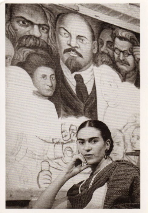 Frida Kahlo sentada frente a un mural de Diego Rivera 