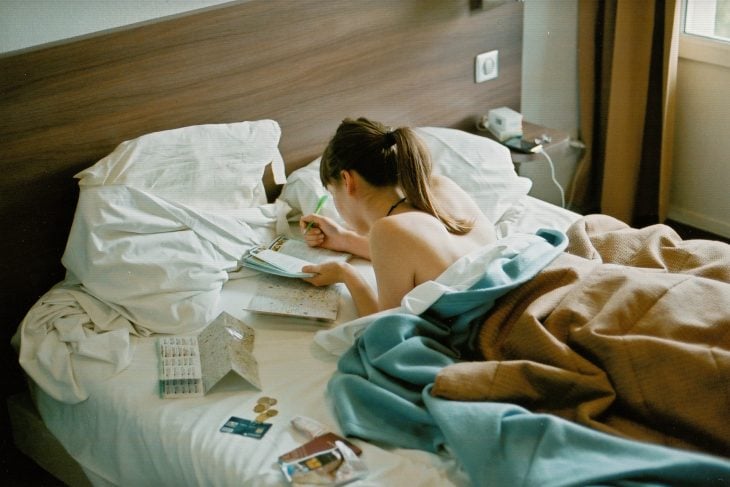 Chica escribiendo en una libreta mientras está recostada boca abajo en su cama 