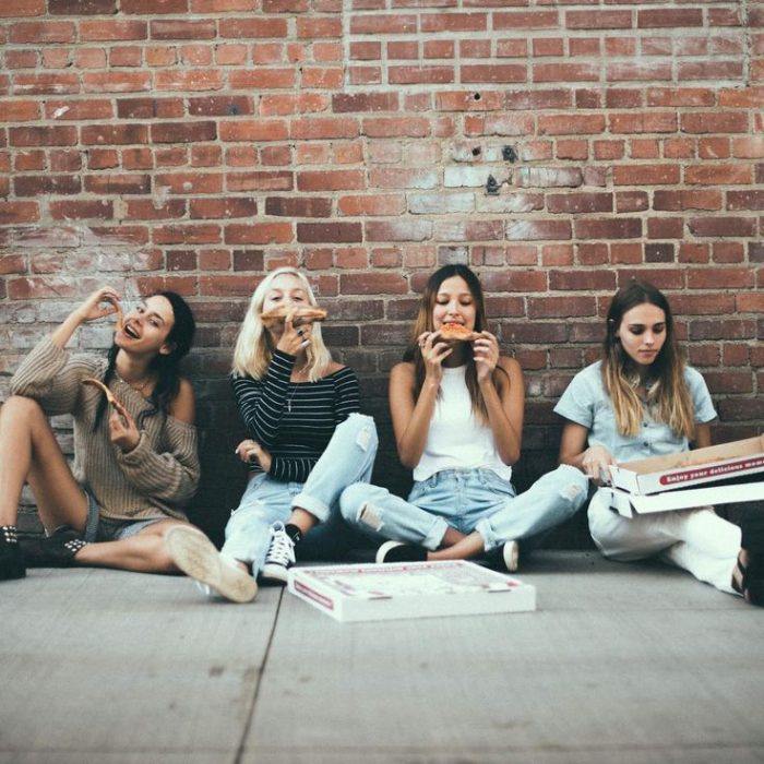 Chicas sentadas en la banqueta y recargadas den la pared comiendo pizza 