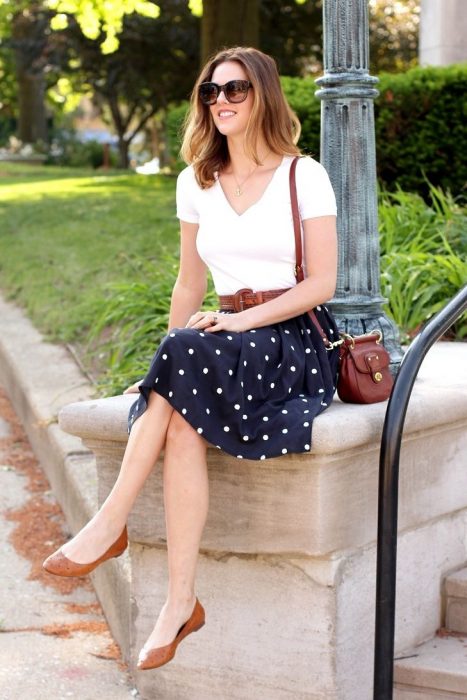 Chica sentada sobre unas escaleras usando una falda azul con puntos y una blusa blanca y flats color café 