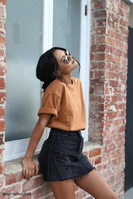 Chica morena con cabello recogido en un chongo bajo posa recargando sus manos en una ventana trasera mientras viste falda negra, blusa naranja y lentes de sol