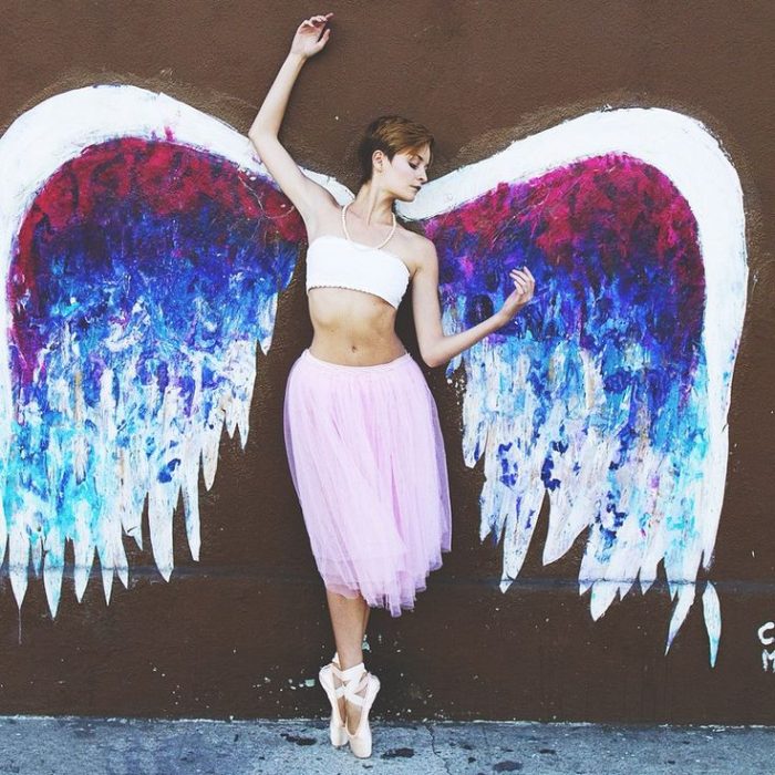 Bailarina de ballet recargada sobre una pared en la que tiene unas alas pintadas 
