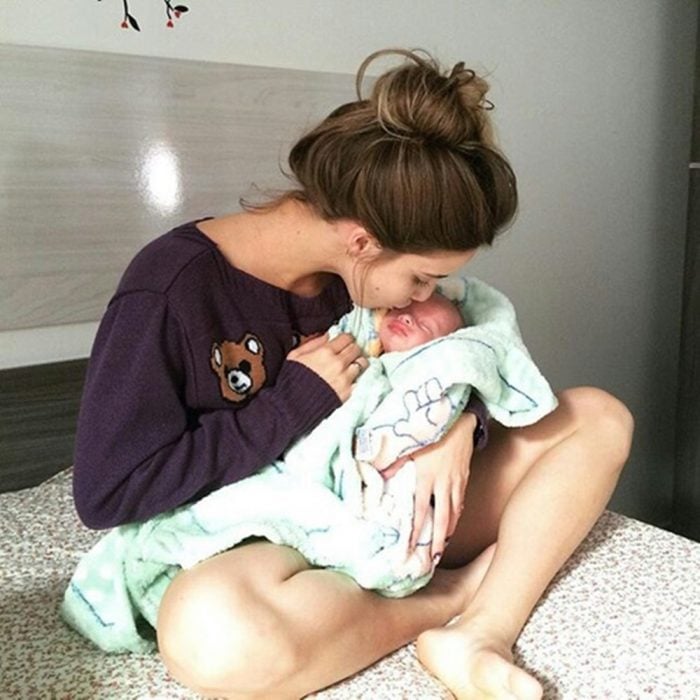 Chica sentada en la cama besando a un bebé recién nacido en la frente 