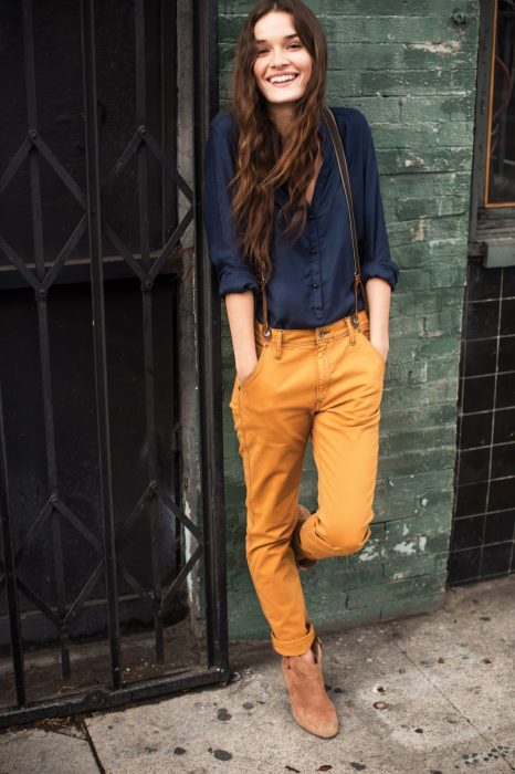 Chica usando una camisa color azul y un pantalón color amarillo mientras está recargada en una pared 