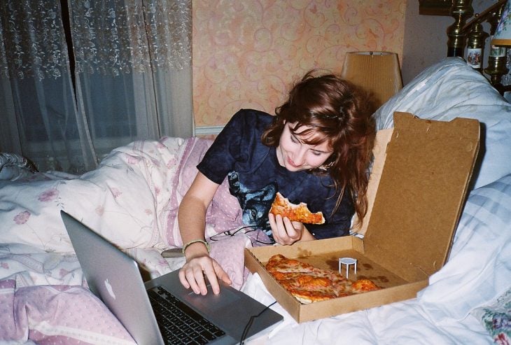 Chica recostada en la cama viendo la computadora y comiendo pizza