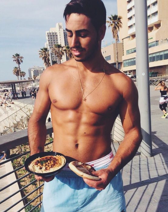 hombre sin playera con comida en la mano y playa atras 