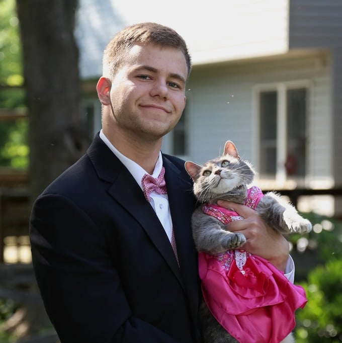 Chico que llevó a su gato al baile de graduación sosteniéndolo mientras el gato usa un vestido rosa 