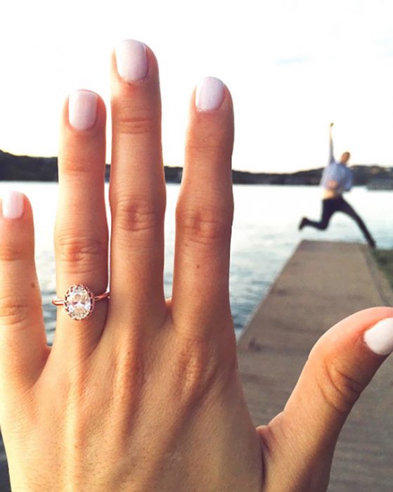 Chica mostrando su anillo de compromiso mientras que su novio salta al fondo de un muelle 