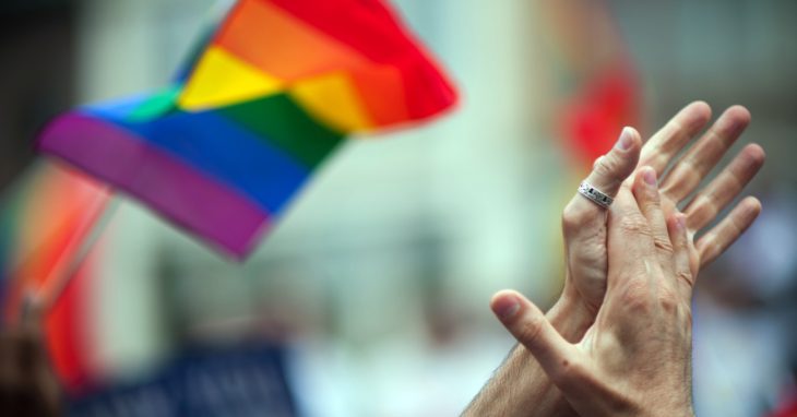 manos entrelazadas con bandera gay al fondo