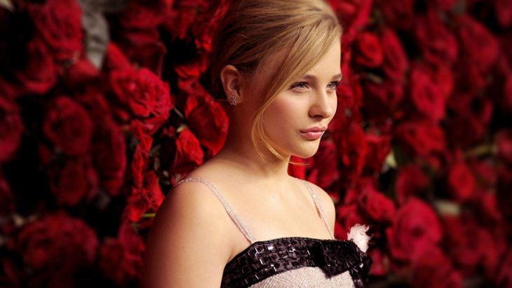 mujer rubia con pared de flores atrás mirando al frente