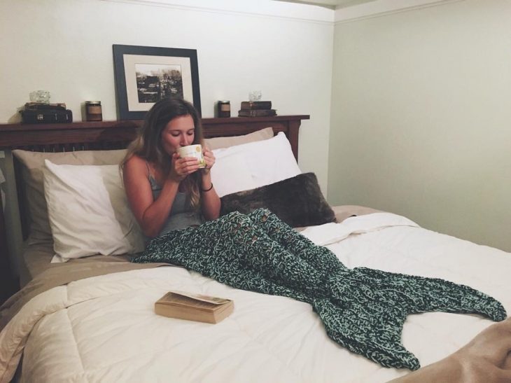 Chica bebiendo una taza de café mientras tiene los pies cubiertos con una frazada de cola de sirena 