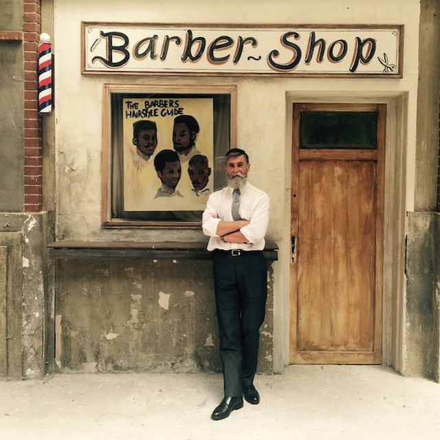 Hombre de 60 años que es modelo posado para una sesión de fotos mientras está recargado en la pared de una barbería 