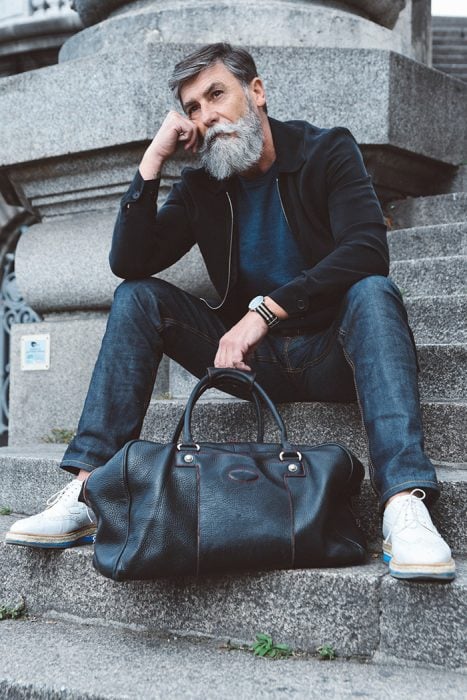 Hombre de 60 años que es modelo posando para una sesión de fotos que incluye una bolsa mientras estpa sentado en unas escaleras 