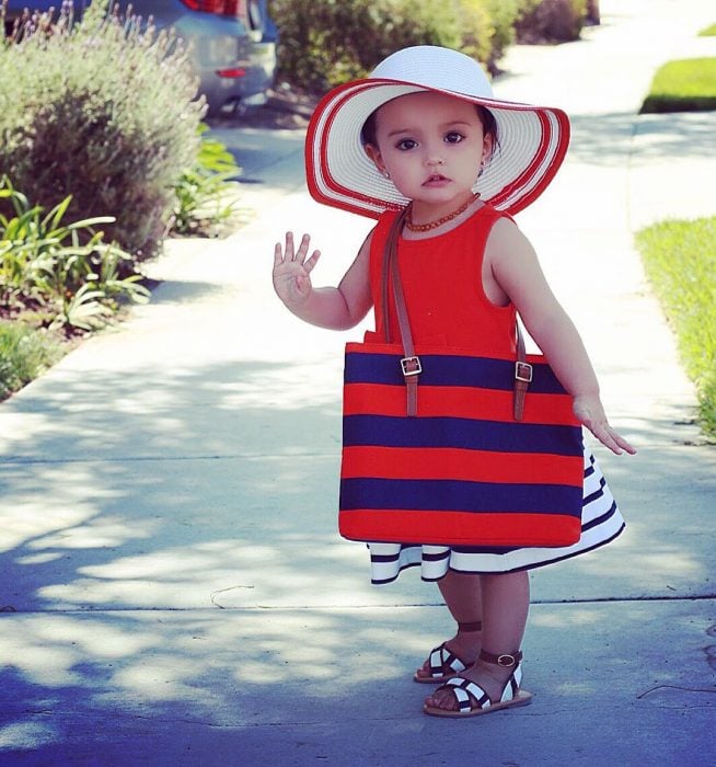 Niña mini fashionista usando un vestido color rojo, bolso de rayas azules y rojas y sombrero blanco 