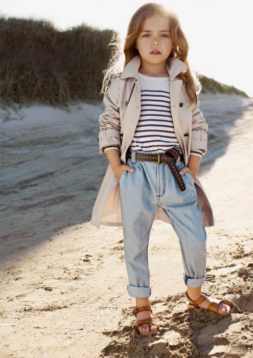 Niña fashionista vestida con una chaqueta, pantalón de mezclilla y sandalias mientras está parada frente al mar 