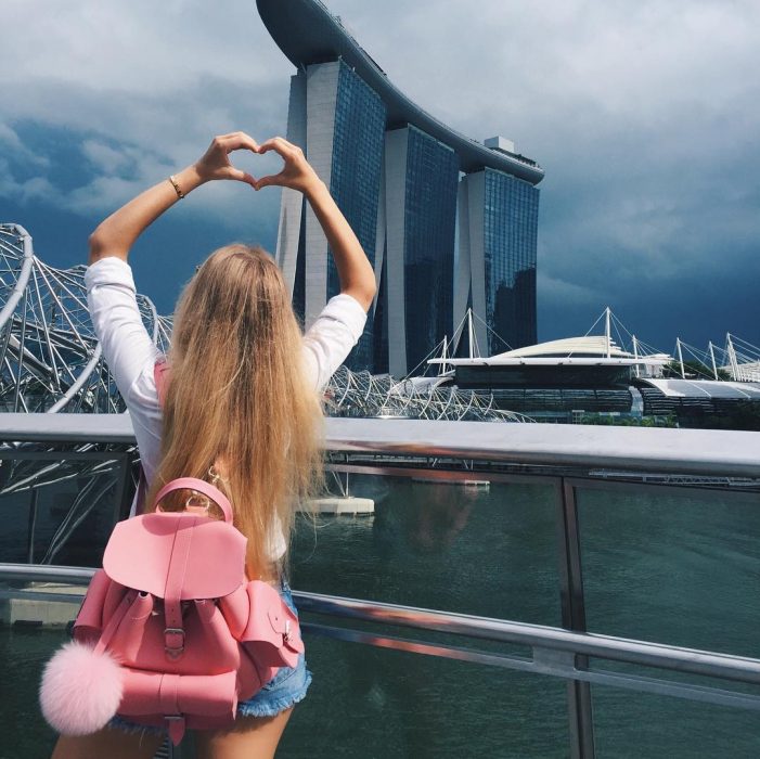 mujer rubia con mochila rosa en cuidad hace corazon con manos 