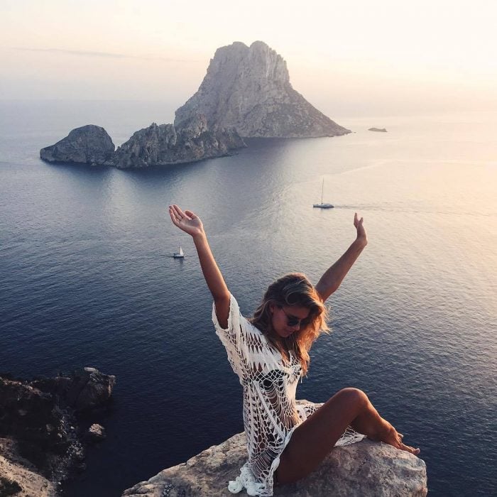 Chica con los brazos levantados mientras está sentada en una piedra admirando el océano 