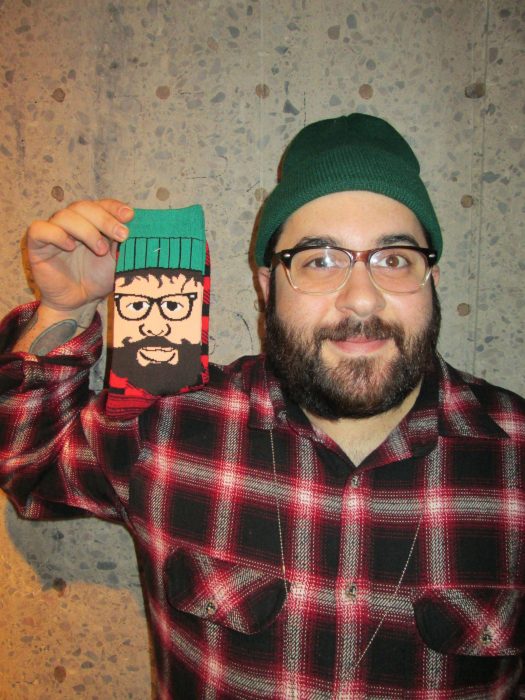 hombre con barba lentes, gorra verde y camisa de leñador sosteniendo celular