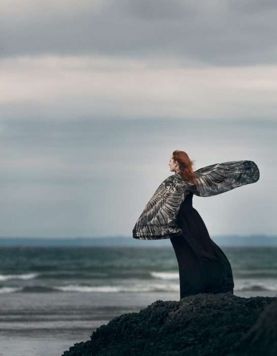 Chica parada sobre una roca contemplando el mar mientras tiene los brazos extendidos 