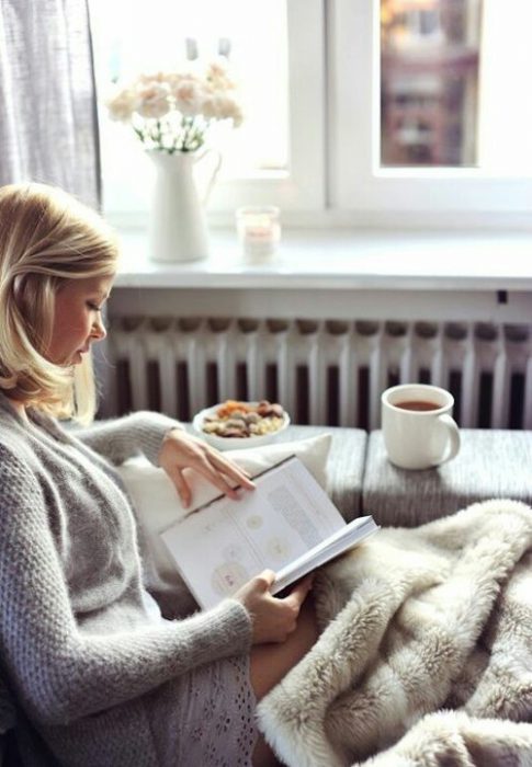 Chica sentada en un sofá leyendo un libro y bebiendo café