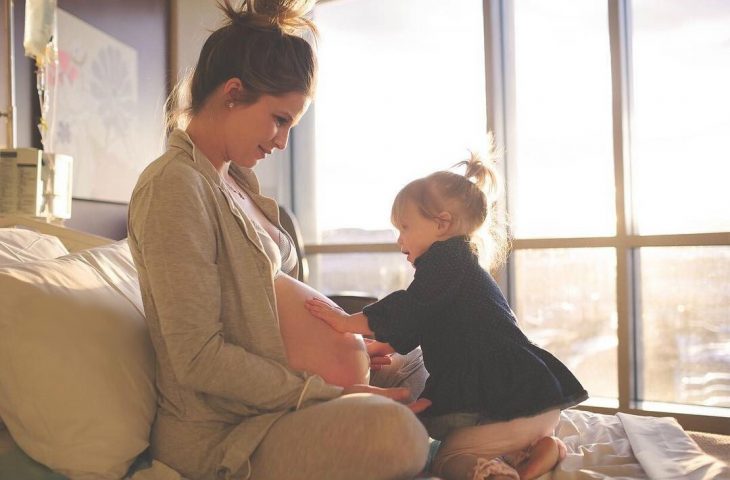 mujer embarazada sentada arriba de cama y niña rubia 