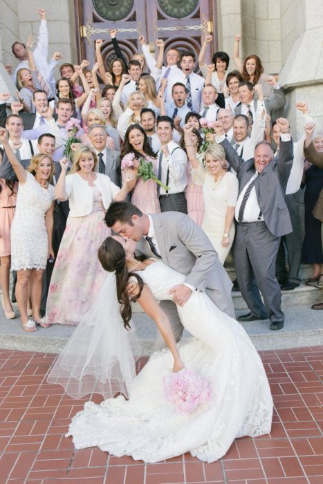 Pareja el día de su boda besándose mientras sus familias festejan atrás de ellos 