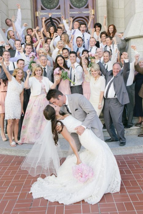 Pareja el día de su boda besándose mientras sus familias festejan atrás de ellos 
