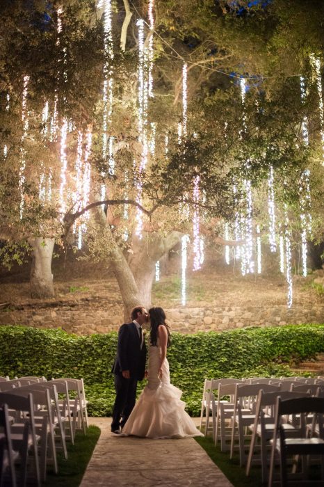 Pareja de novios besándose bajo un árbol cubierto de luces