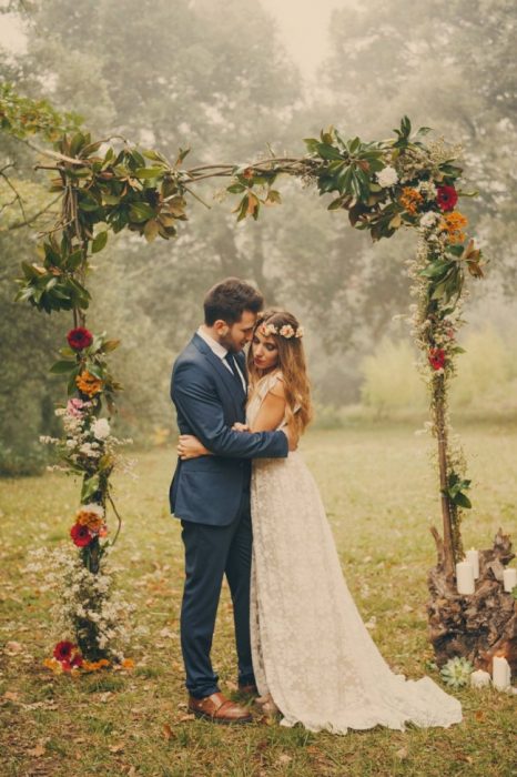 Pareja de novios con una boda boho parados bajo un marco de madera con flores
