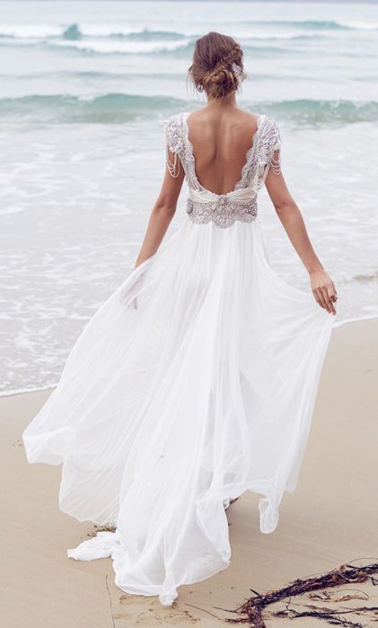 Chica con un vestido de novia con escote en la espalda caminando por la playa 