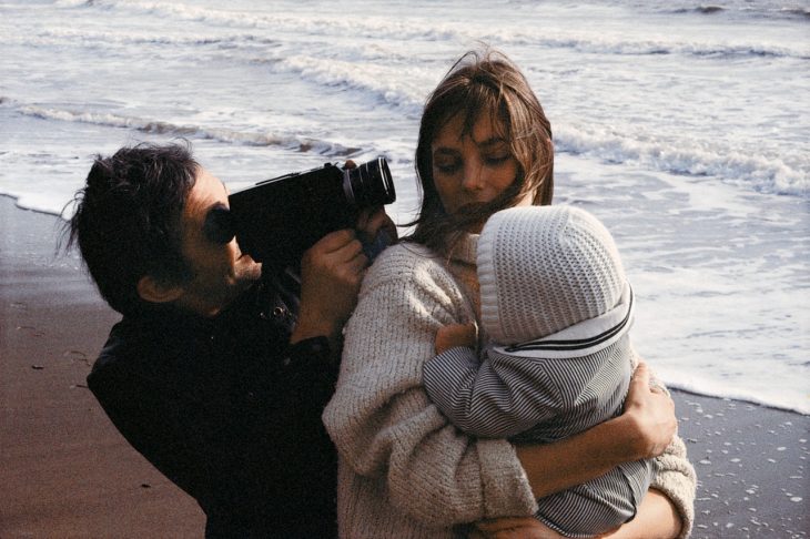 hombre grabando a su mujer cargando un bebé en la playa 