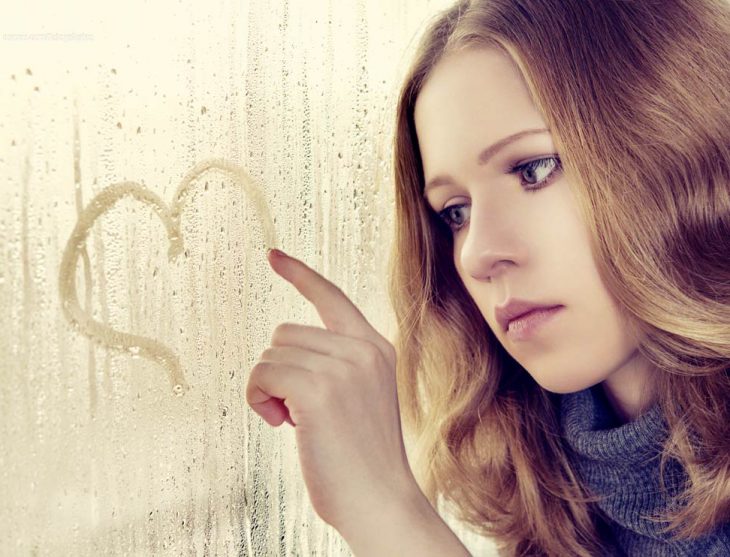 Chica mirando por un vidrio mientras dibuja un corazón. 