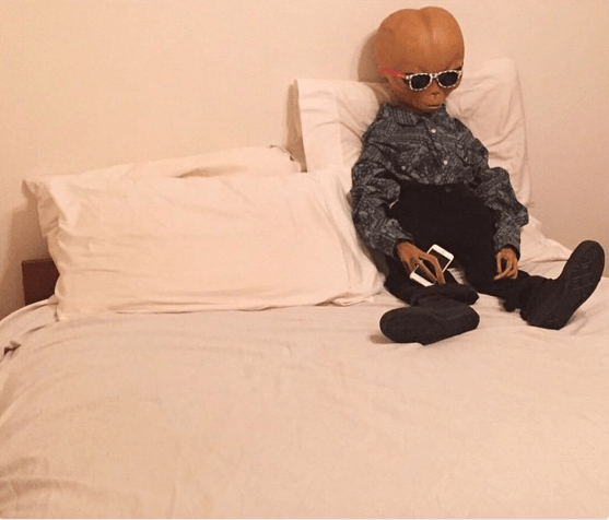 Extraterrestre sentando en una cama con un celular en la mano. 