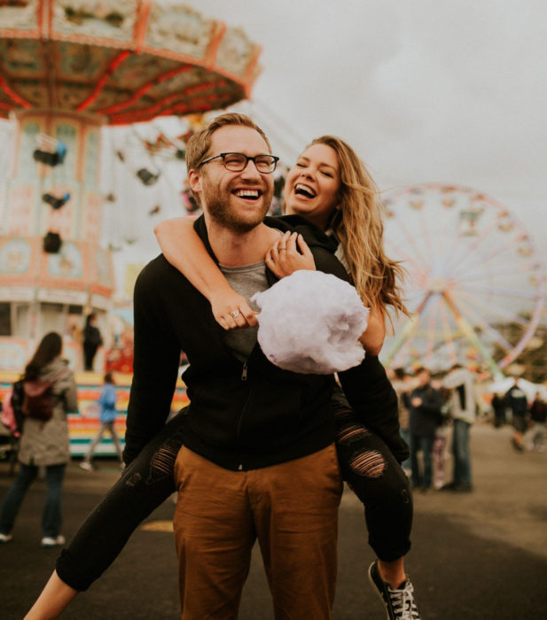 Pareja de novios riendo en parque de diversiones, chico cargando a chica en su espalda