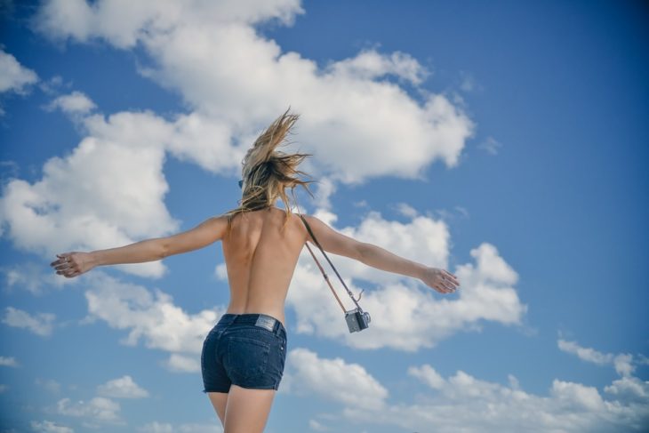 mejer feliz sin playera frente a las nubes y con una camara