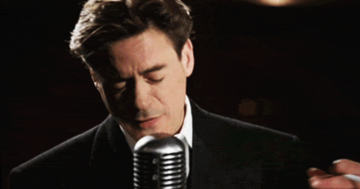 Hombre usando traje sastre negro, cantando frente a un micrófono antiguo plateado, con los ojos cerrados, Robert Downey Jr. 