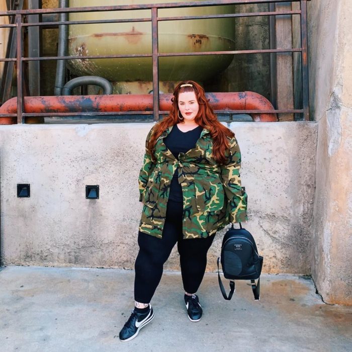 Tess Holliday posando con jeans, zapatos y blusa larga negras con una chaqueta militar 