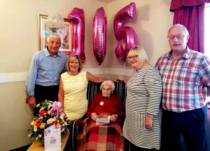 hombres y mujeres alrededor de anciana con globos de cumpleaños rosa 