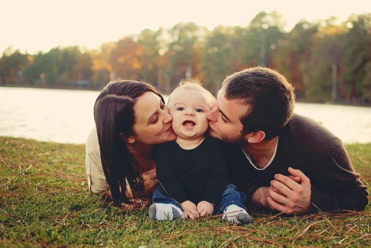 Familia posando para foto en el pasto; mamá y papá dando beso en el cachete a su bebé sonriente