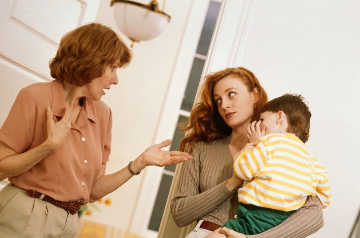 Relación conflictiva con la suegra; mujer mayor mirando a su nuera; chica pelirroja cargando a su bebé