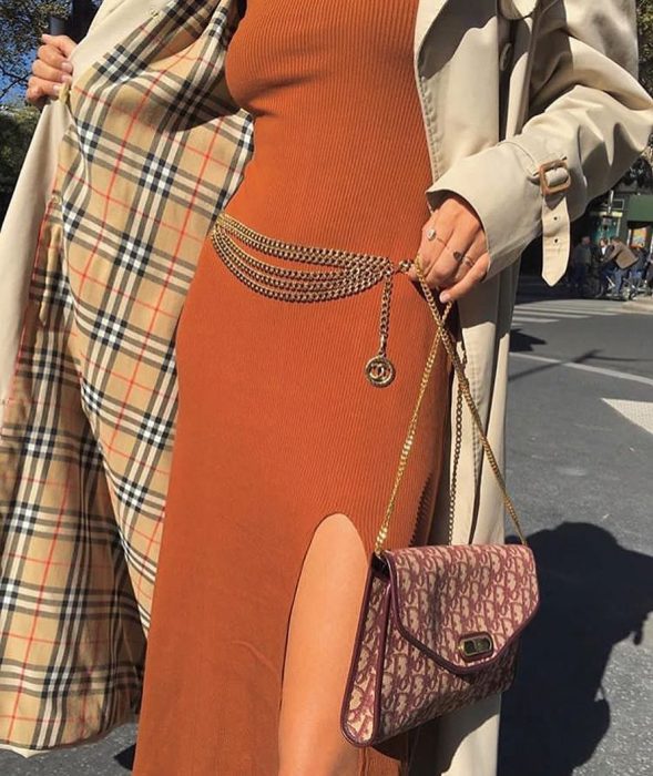 mujer con vestido largo naranja y abrigo beige con cinto de cadena