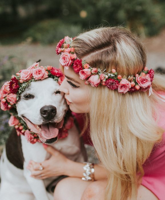 Chica rubia con corona de flores abrazando a un perro sonriente con corona de flores