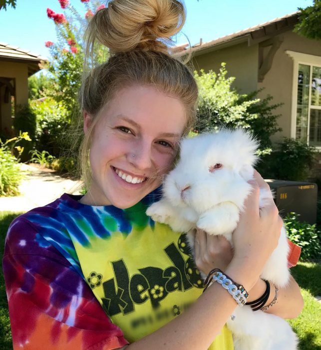 Chica de cabello rubio con un chongo y playera de colores abrazando a un conejo blanco