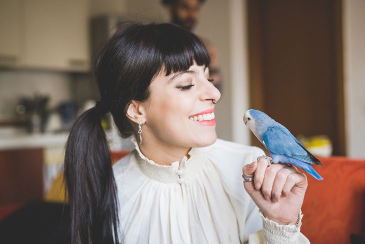 Chica de cabello largo agarrado en una coleta y blusa blanca sosteniendo a un perico azul