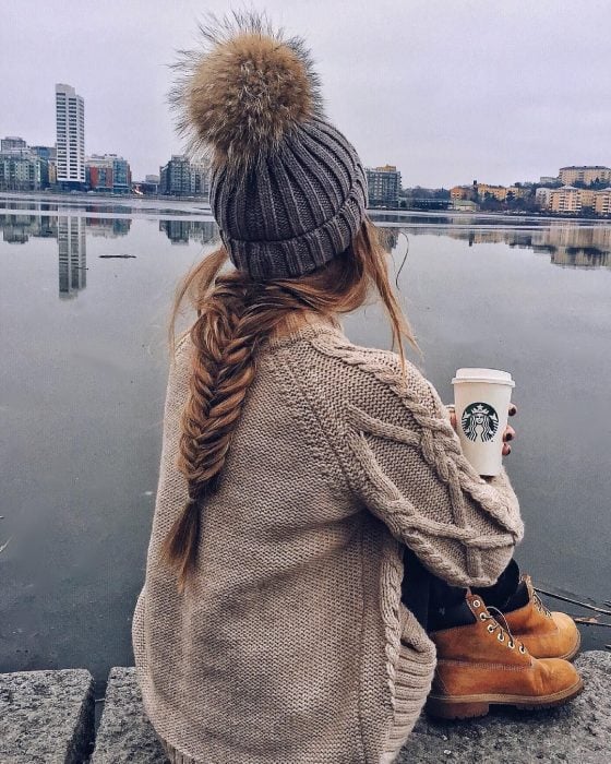 Chica de cabello largo y trenzado usando gorro de invierno sentada y bebiendo café