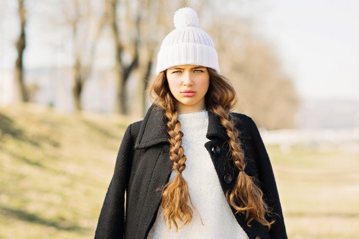 Chica con cabello largo y trenzado vistiendo un gorro blanco d e invierno y un abrigo negro