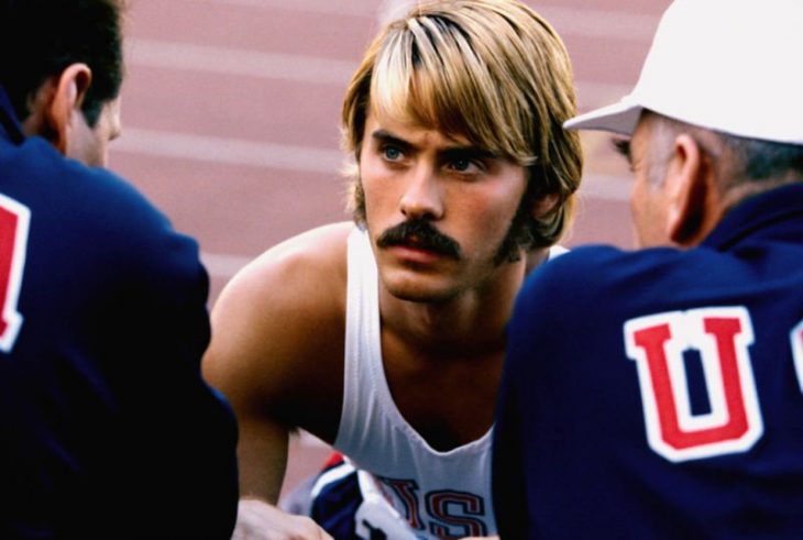 Hombre rubio y de bigote en una pista para correr mirando a otros dos hombres