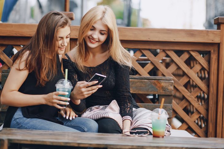 Par de amigas con blusa negra sentadas en una banca tomando una bebida y mirando el celular