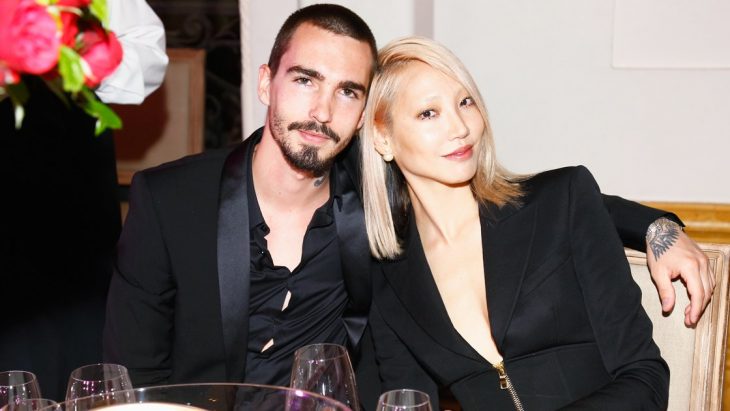 Soo Joo Park y Jack Waterlot, pareja abrazada, mujer asiática rubia con traje negro y hombre con barba y traje negro